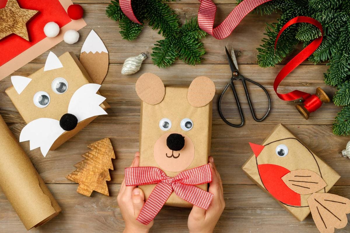 Christmas-themed crafts and gift wrapping on a wooden surface. Presents are decorated with animal faces, using paper and googly eyes. Foliage, ribbons, and scissors are also present. A pair of hands holds a bear-themed gift.