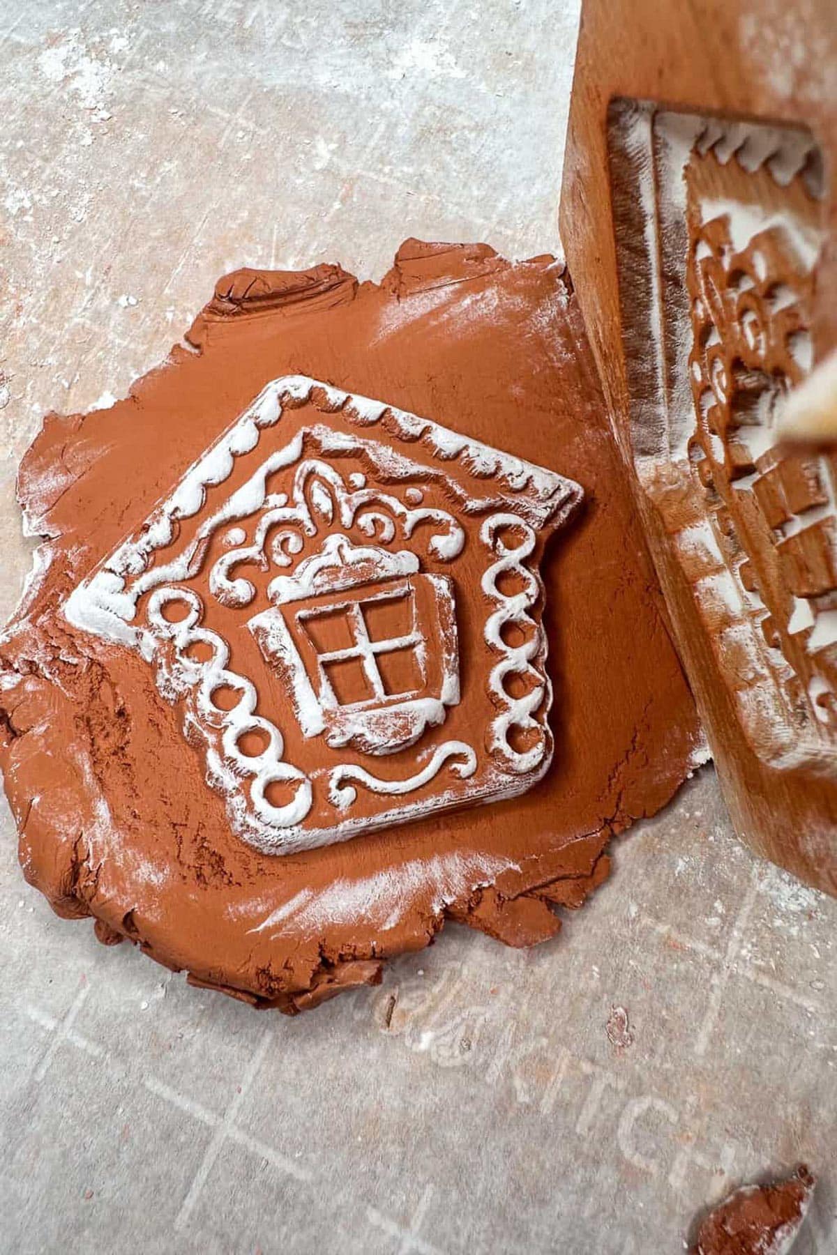 Close-up of a clay imprint featuring an ornate, house-shaped design with a window and decorative patterns. The imprint is on a flat piece of clay, next to the stamp used to create it.