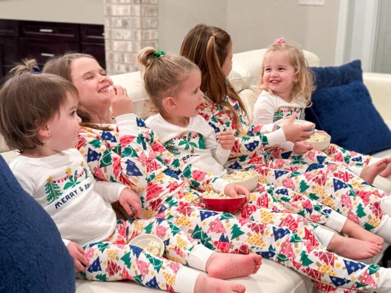 Five children wearing colorful holiday pajamas sit on a white couch, laughing and eating popcorn. They appear joyful and festive, surrounded by large blue pillows.