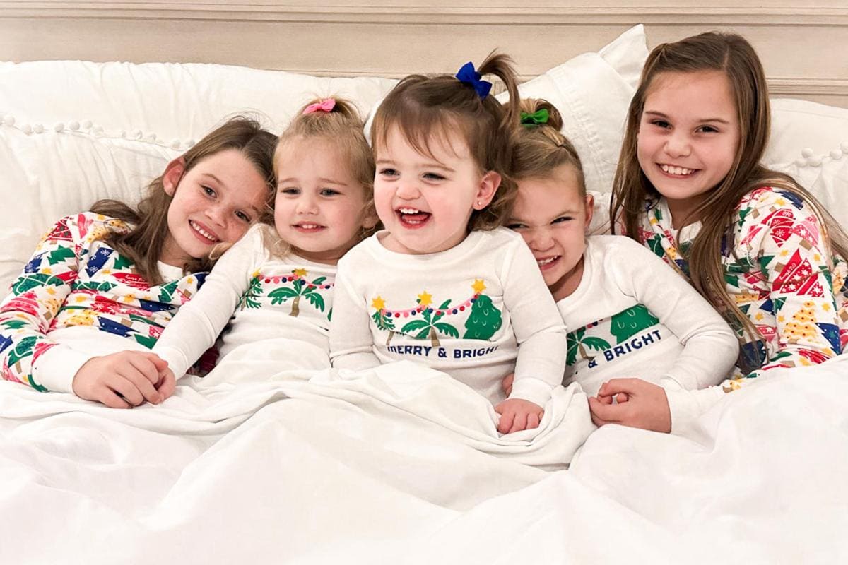 Five young children in festive pajamas are snuggled together on a bed. The room has a cozy and cheerful atmosphere. The pajamas feature colorful holiday patterns. The children are smiling and looking happy.