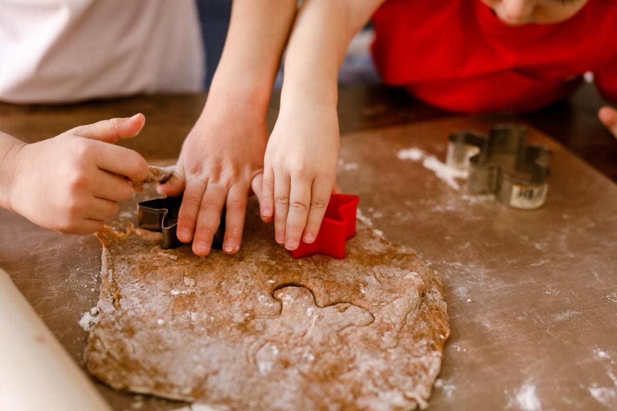 Two people use star-shaped cookie cutters on rolled-out dough. One cutter is red, the other is grey. The dough is on a floured brown surface, and a rolling pin is partially visible.