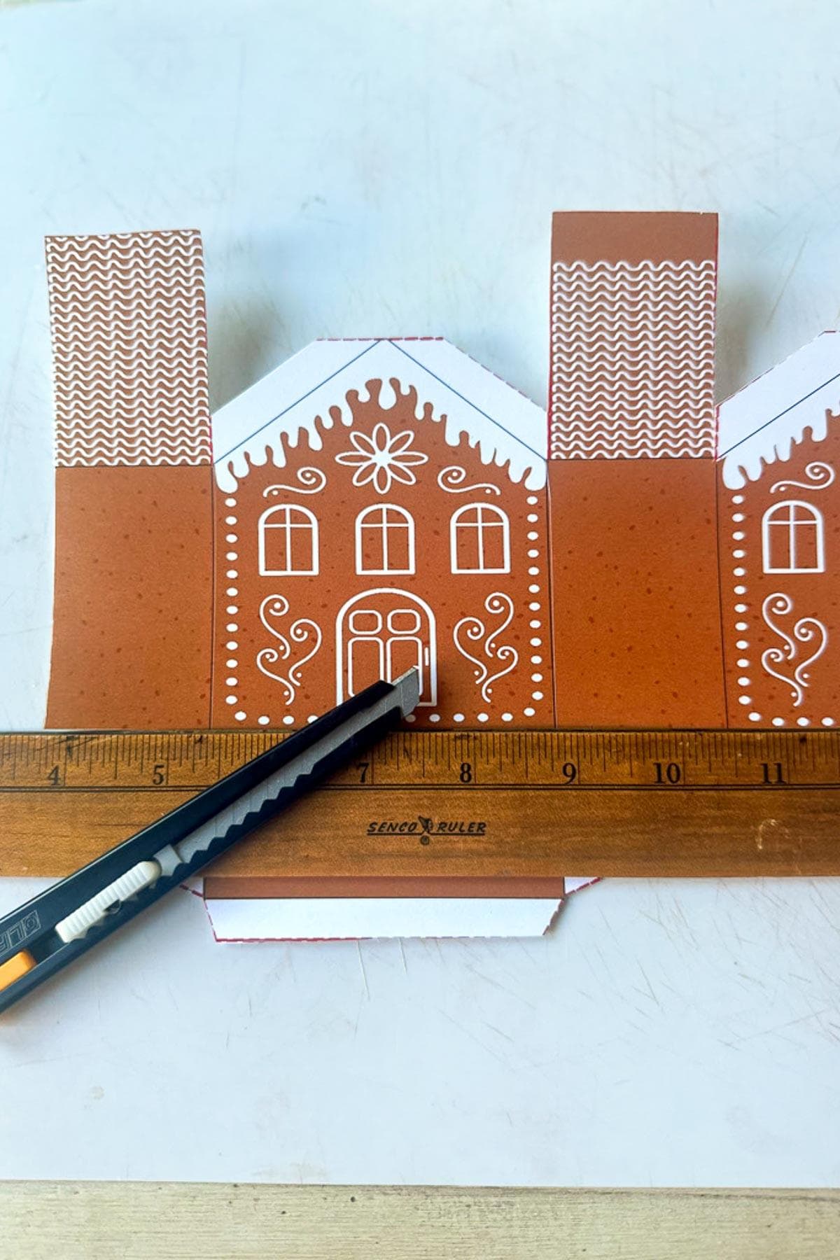 A gingerbread house template made of paper is on a table. A ruler and a craft knife are placed on top, showing a precise cutting line. The template features drawn-on doors, windows, and decorative patterns.