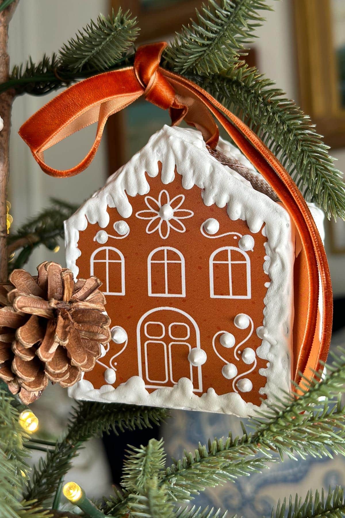 A gingerbread house ornament decorated with white icing designs hangs on a Christmas tree branch. An orange ribbon is attached to the ornament, and a pinecone is nearby. The background is softly blurred.