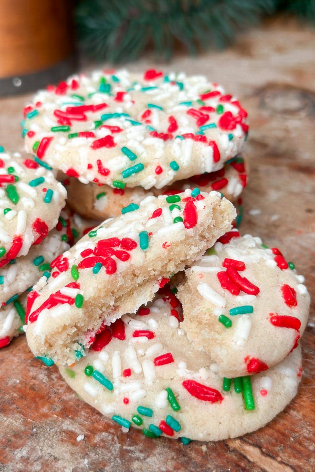 A stack of festive cookies topped with red, green, and white sprinkles sit on a wooden surface. One cookie is broken in half, revealing a soft interior. A hint of greenery is visible in the background.