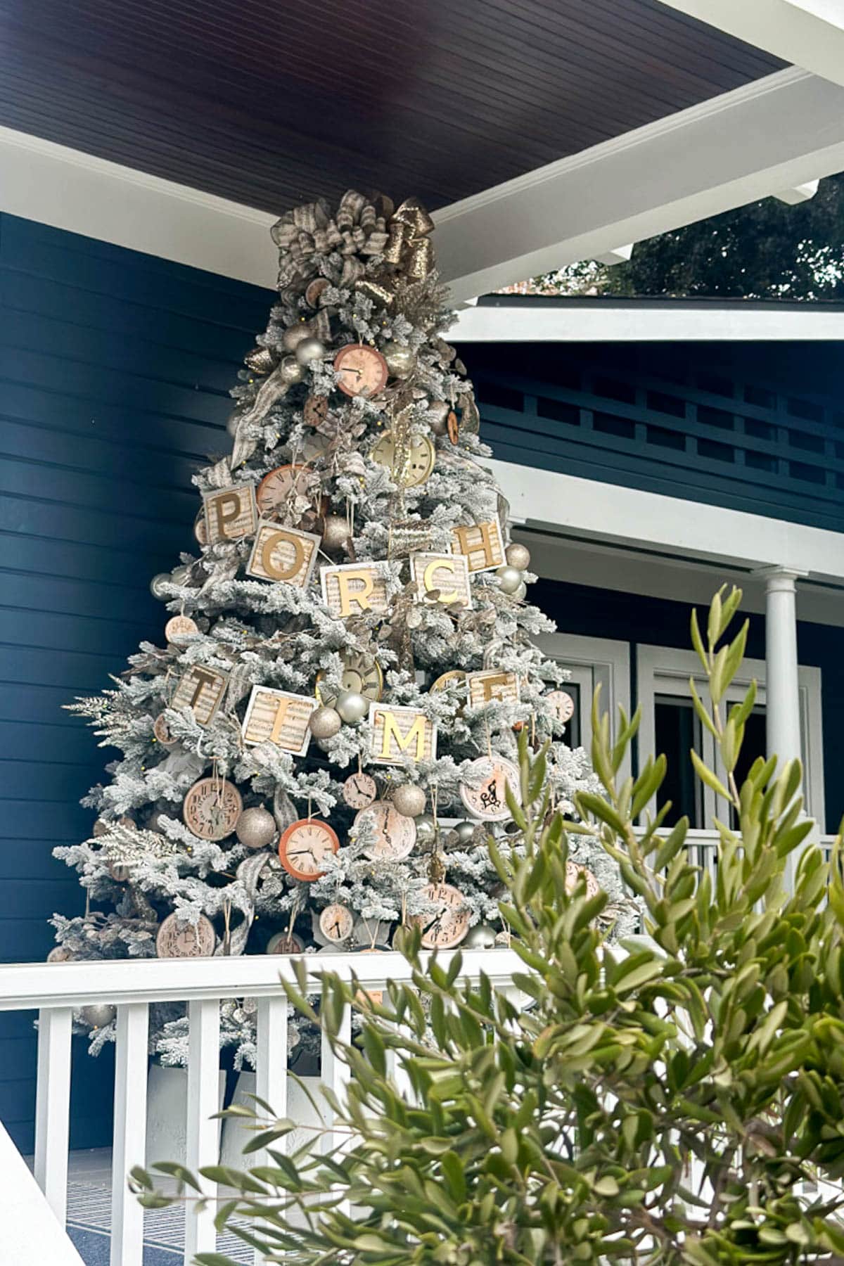 A frosted Christmas tree stands on a porch, adorned with large letters spelling “Porch Time,” gold baubles, and silver DIY vintage Christmas ornaments. The tree is set against a blue house with white railings, while green foliage adds charm in the foreground.