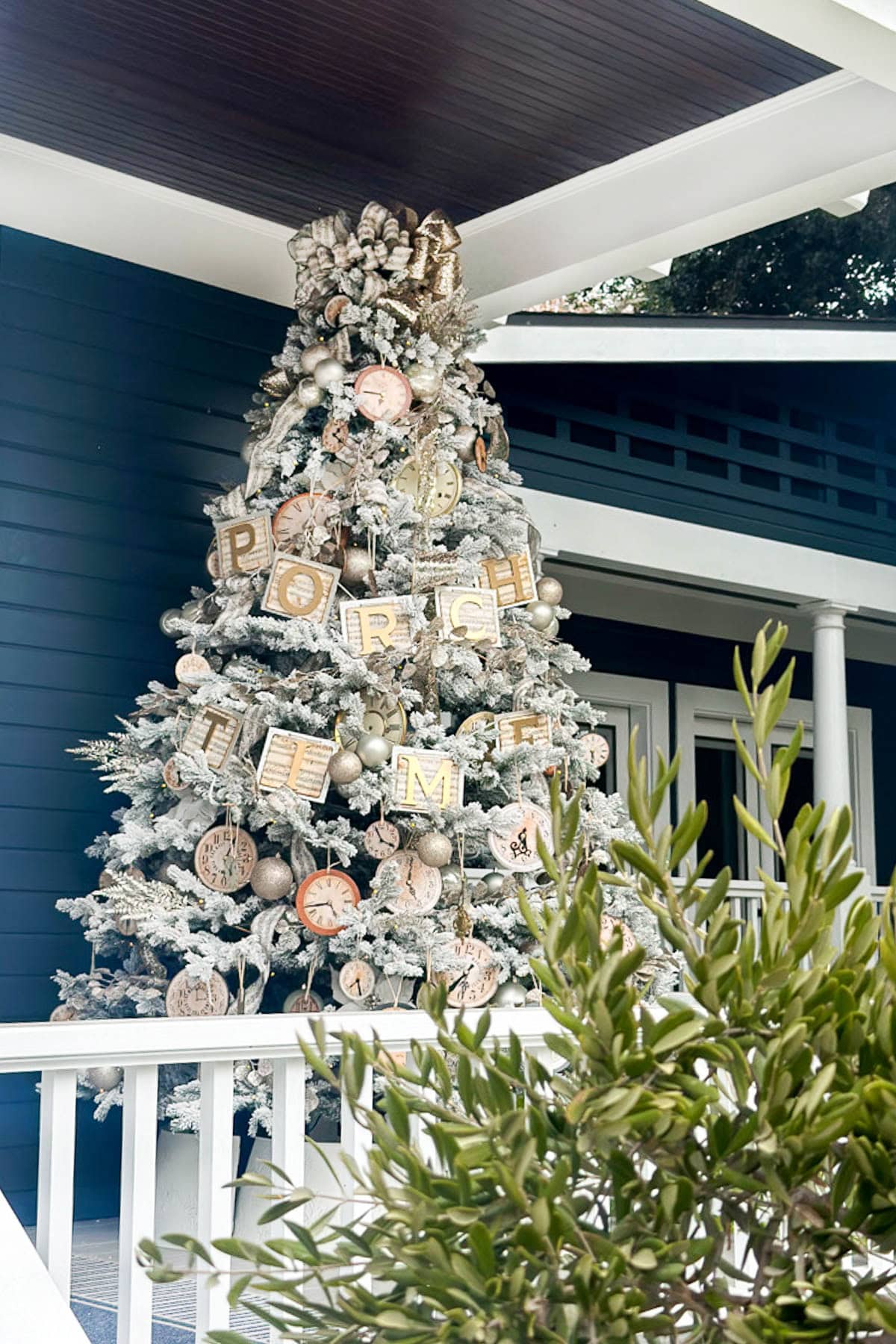 A snow-covered Christmas tree, adorned with DIY vintage Christmas ornaments and clock faces in gold, stands proudly on the porch of a dark blue house. A garland spelling "PORCH TIME" adds charm, while greenery from a nearby bush peeks through in the foreground.