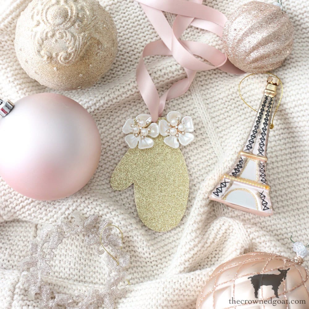 Festive ornaments on a textured knit background, including a glittery mitten with floral detail, pink ribbon, a ball ornament, a decorative Eiffel Tower, and a shiny crown.