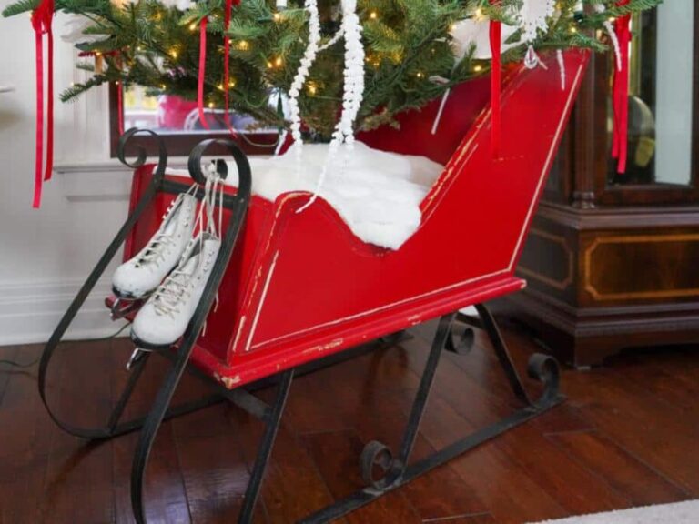 A red sleigh with black runners serves as a festive display indoors, nestled on an artificial Christmas tree stand. Filled with fake snow and positioned under a glowing tree, a pair of white ice skates hangs gracefully on the front.