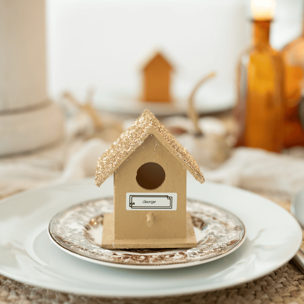 A small, decorative birdhouse with a glittery roof sits on a floral-patterned plate. It has a label reading "George." The setting is on a table with warm-toned decor, including a lit candle and amber glass bottle in the background.