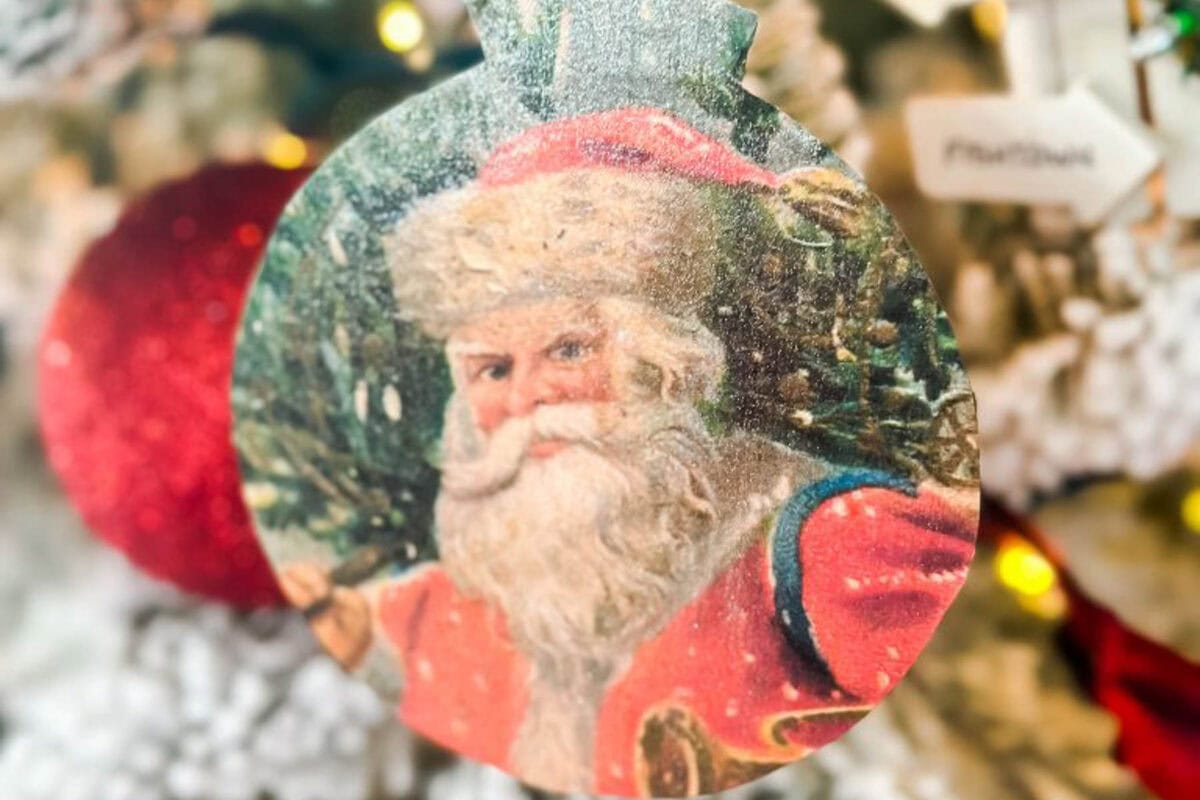 Close-up of a DIY vintage Christmas ornament featuring a charming illustration of Santa Claus with his classic red suit and white beard. In the background, soft, blurred lights dance among snowy branches, capturing the nostalgic spirit of the season.