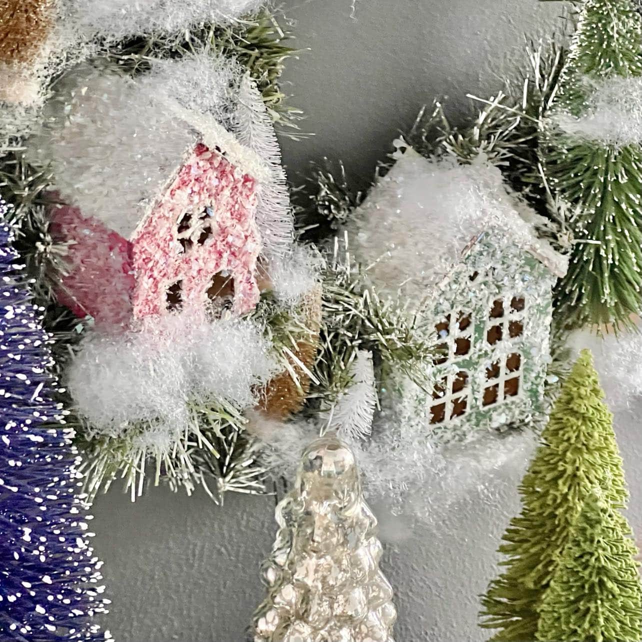 Close-up of miniature snowy houses in pastel pink and green surrounded by artificial pine trees and snow. The scene has a festive, wintery feel with various shades of green adding depth to the decoration.