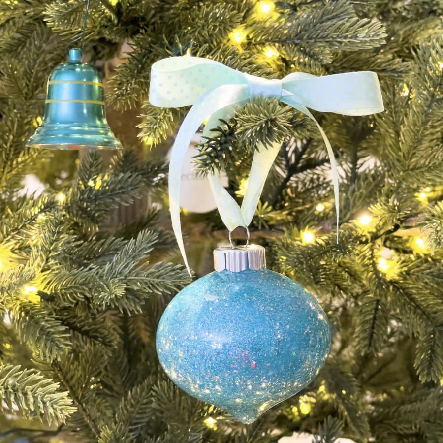 A glittery blue ornament with a light blue ribbon hangs on a Christmas tree, surrounded by twinkling lights and green branches. Nearby, a green bell-shaped ornament is visible.