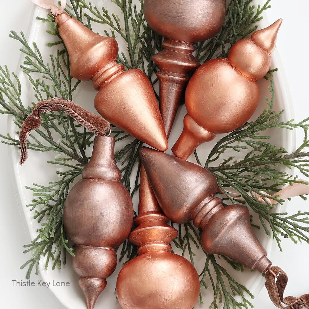 A collection of copper-colored, uniquely shaped ornaments arranged on a bed of evergreen branches on a white surface. The décor includes round and pointed designs with a metallic finish.