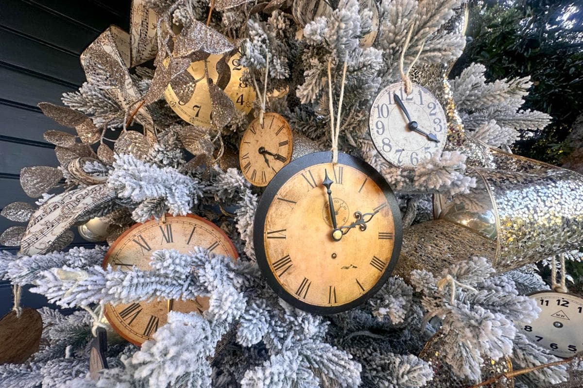 A Christmas tree decorated with various vintage clocks and glittery ornaments. The tree is dusted with faux snow, creating a wintry effect. Each clock shows a different time, adding a whimsical touch to the festive display.