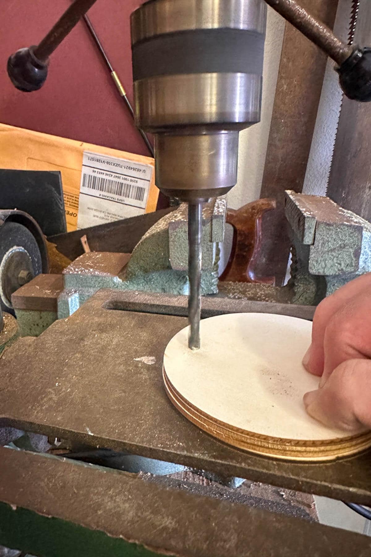 A person uses a drill press to bore a hole through a stack of round wooden pieces. The drill bit is centered on the stack, and a vise secures the wooden pieces in place on the workbench.