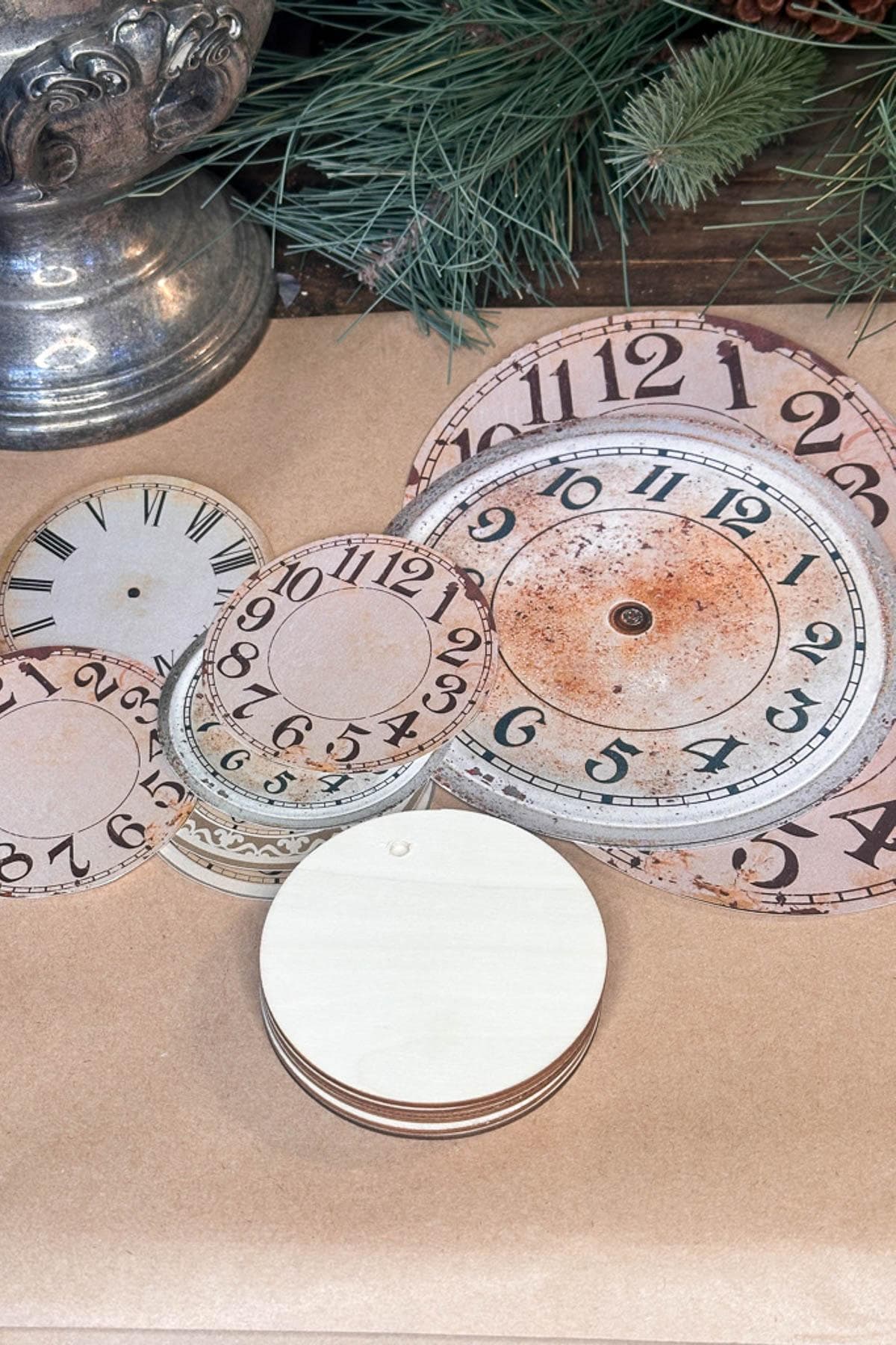 A collection of vintage clock faces in various sizes and designs laid out on a table. There are ornate numerals and weathered patinas. Pine branches and a silver vase are partially visible in the background.