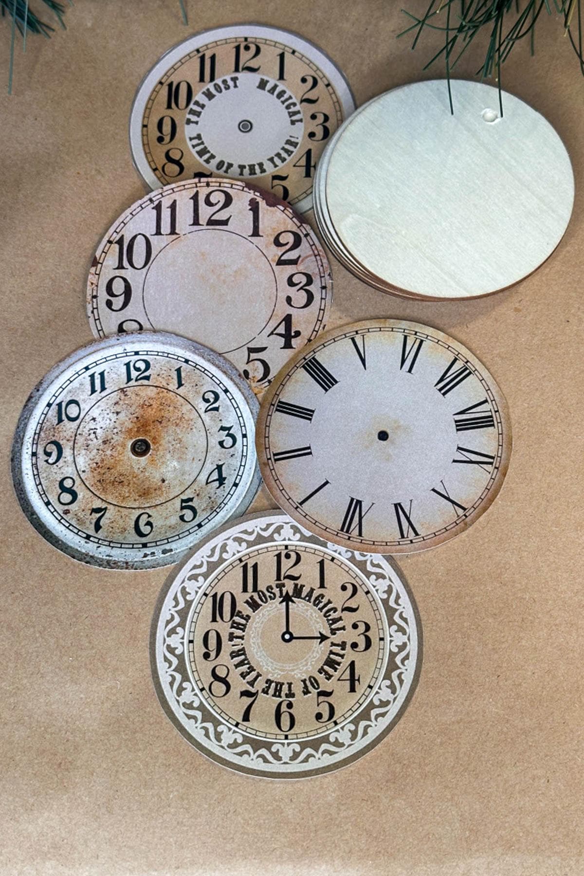 A collection of seven vintage clock face cutouts laid out on a textured brown surface. The designs include Roman and Arabic numerals, some with ornate patterns and weathered finishes.