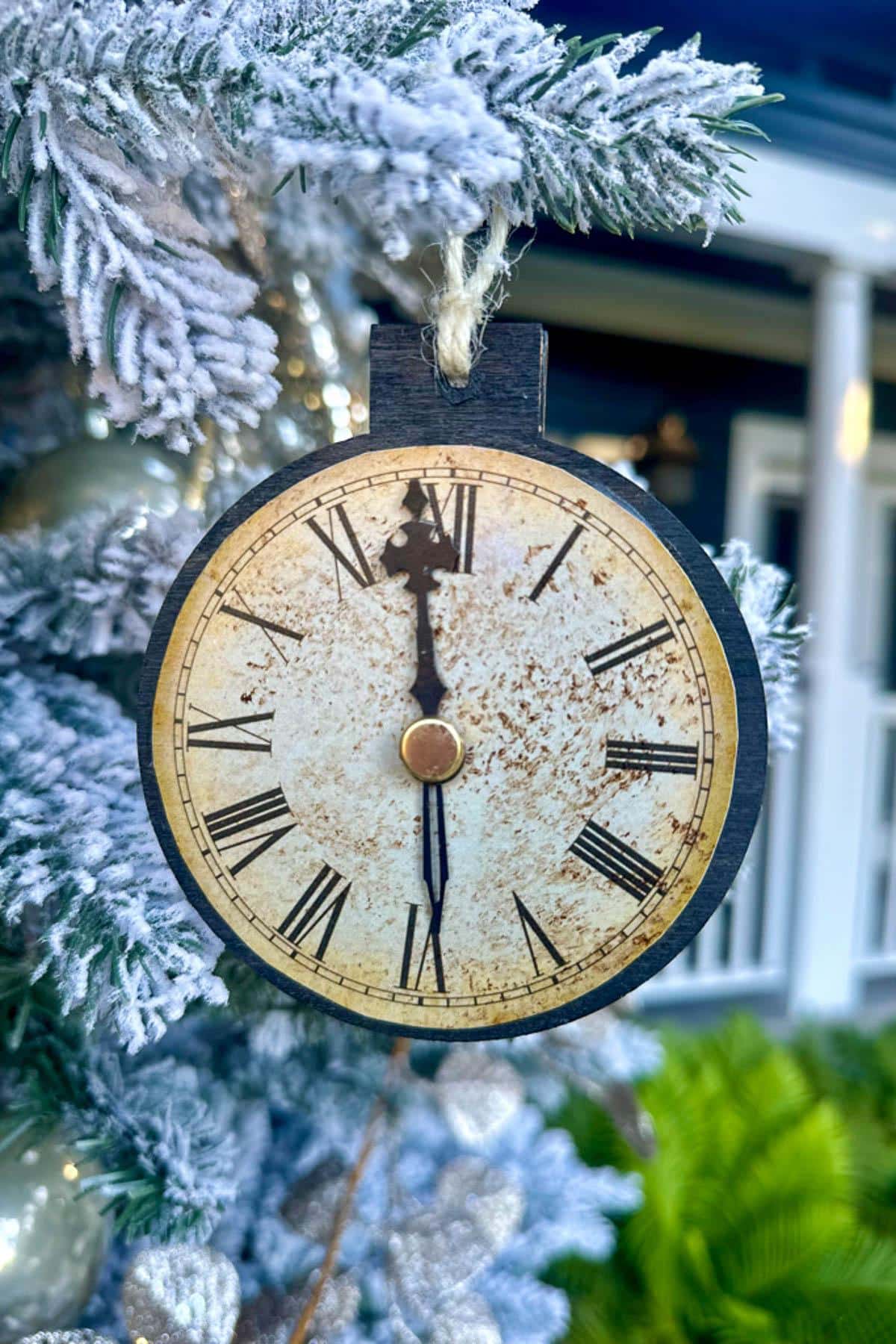 A vintage clock ornament with Roman numerals is hanging on a frosted Christmas tree. The clock hands are near midnight. Snow-covered branches and blurred background suggest a festive, winter setting.