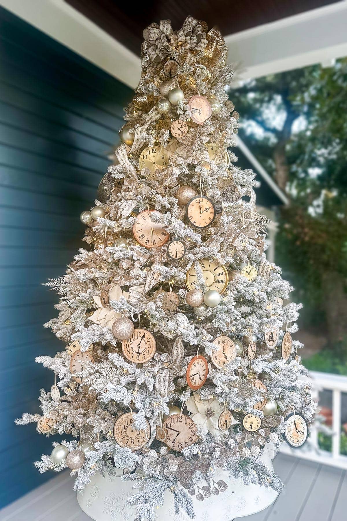 A Christmas tree adorned with vintage-style clock ornaments and metallic baubles, set on a white porch. The tree has frosted branches and a sparkling ribbon on top.