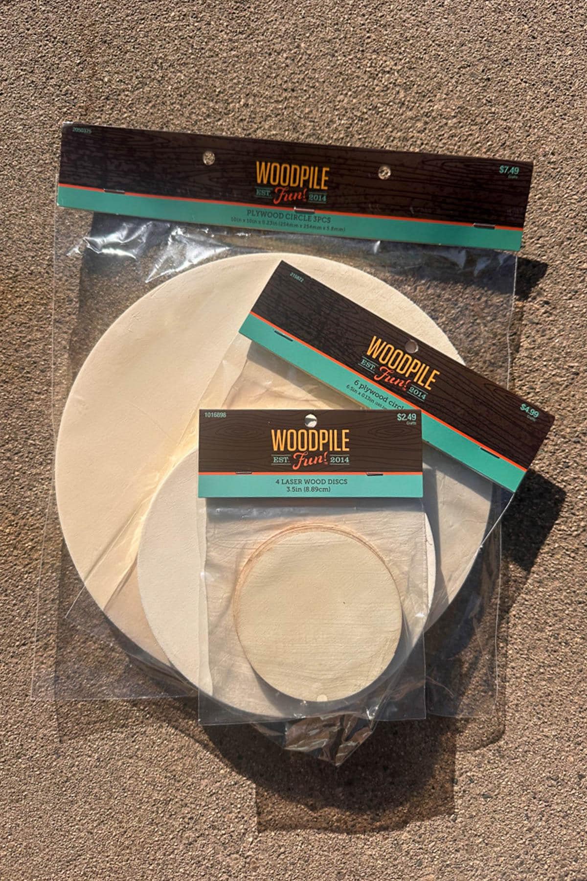 Two packages of wooden circles from Woodpile Fun! are displayed on a textured surface. The larger pack contains multiple sizes of circles, while the smaller pack has four wood rounds, all sealed in clear plastic.