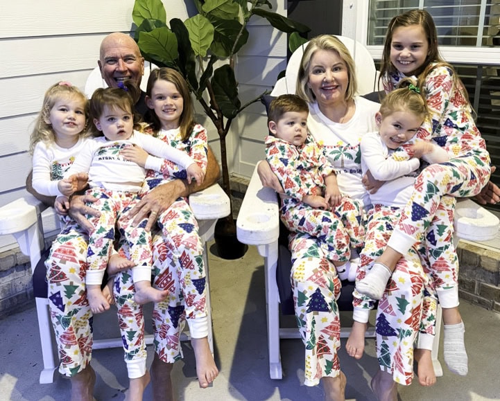 A family wearing matching colorful pajamas stands joyfully in front of a house, capturing the essence of a Christmas Pajama Party. Two adults sit on white chairs surrounded by their six lively grandkids, some perched on their laps while others stand nearby. A large plant adds a touch of greenery to the scene.