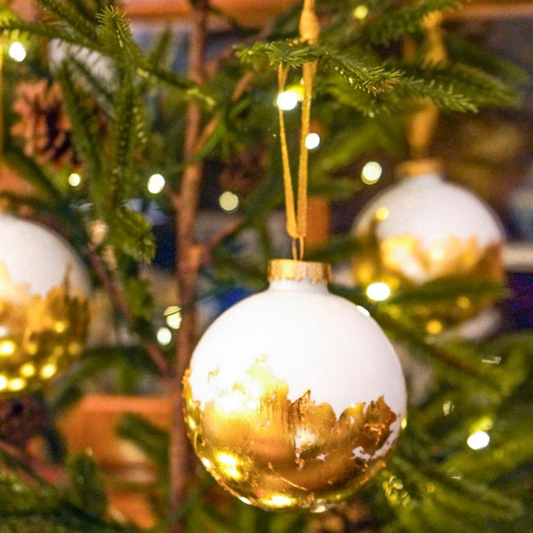 A close-up of a Christmas tree adorned with white and gold baubles. The ornaments have a golden leaf design on the bottom half. Soft white string lights add a warm glow to the scene.