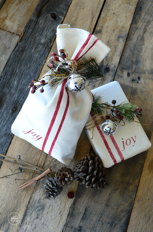 Two wrapped gifts in white fabric with red stripes and the word "joy" are on a wooden surface, exuding the charm of DIY vintage Christmas ornaments. Each is adorned with pinecones, pine branches, red berries, and silver bells. Pinecones are scattered around them.