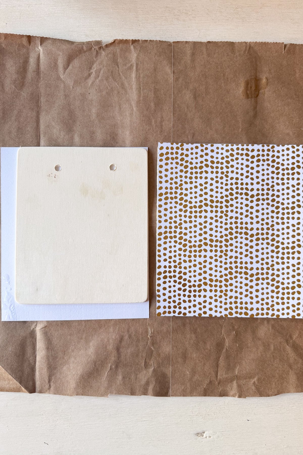 A wooden square with two holes is placed on the left. A patterned sheet of paper with small gold dots on a white background is on the right. Both rest on a crumpled brown paper surface.