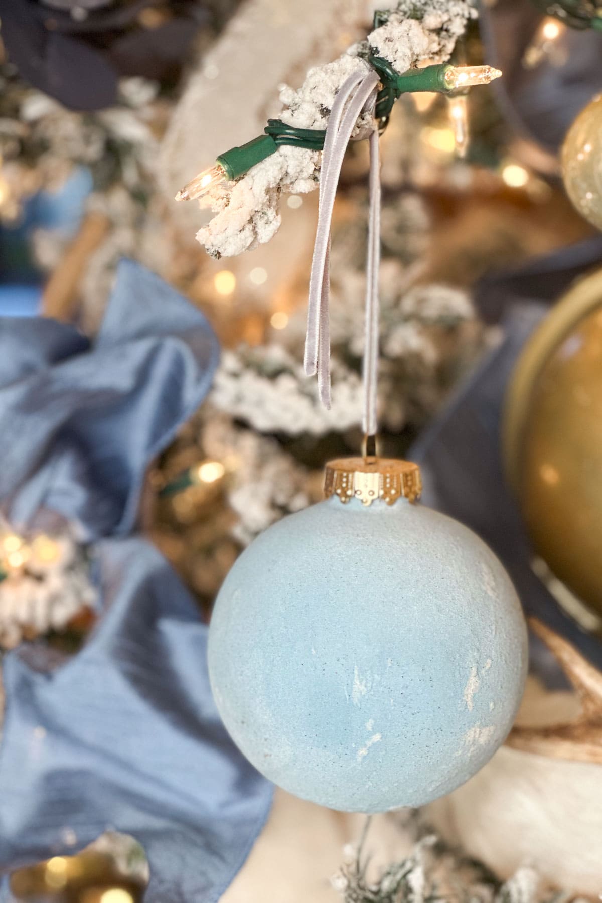 A close-up of a light blue Christmas ornament hanging on a snowy branch decorated with small white lights and a blue ribbon. The ornament is accented by other blurred decorations and lights in the background.