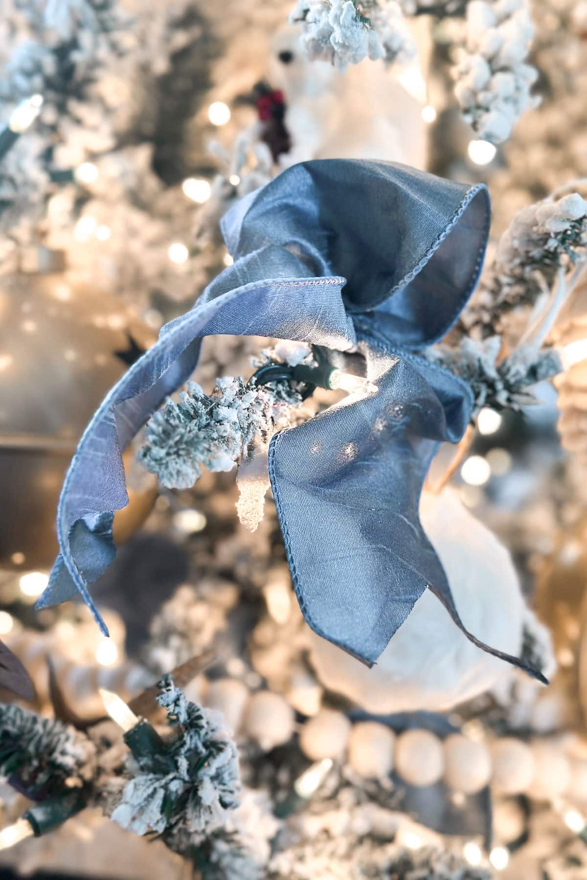 A close-up of a Christmas tree adorned with blue and white Christmas decor, featuring a shimmering blue ribbon and elegant white ornaments. Snow-like frosting covers the branches, while warm white lights cast a festive glow.