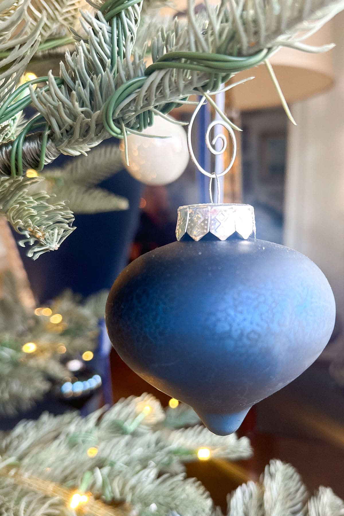 A close-up of a matte blue Christmas ornament hanging from a branch of a decorated tree. The ornament has a metallic cap and curly hook. Soft, warm lights and blurred background elements create a cozy atmosphere.