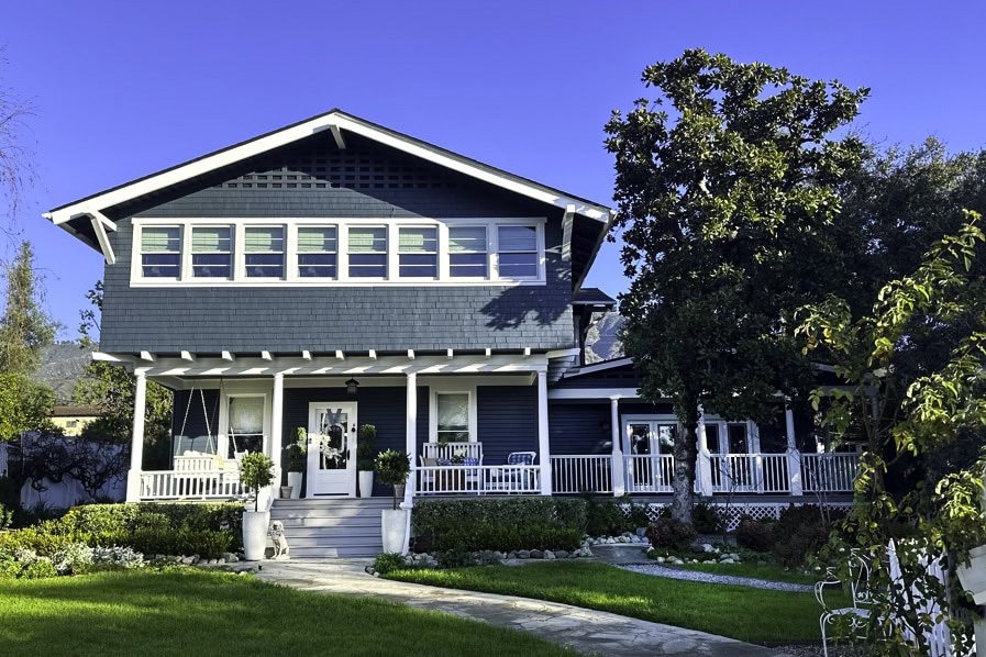 A two-story blue craftsman-style house with white trim, adorned with blue and white Christmas decor, features a wide front porch, swing, and multiple windows. The lush front yard is landscaped with trees and shrubs under a clear blue sky.