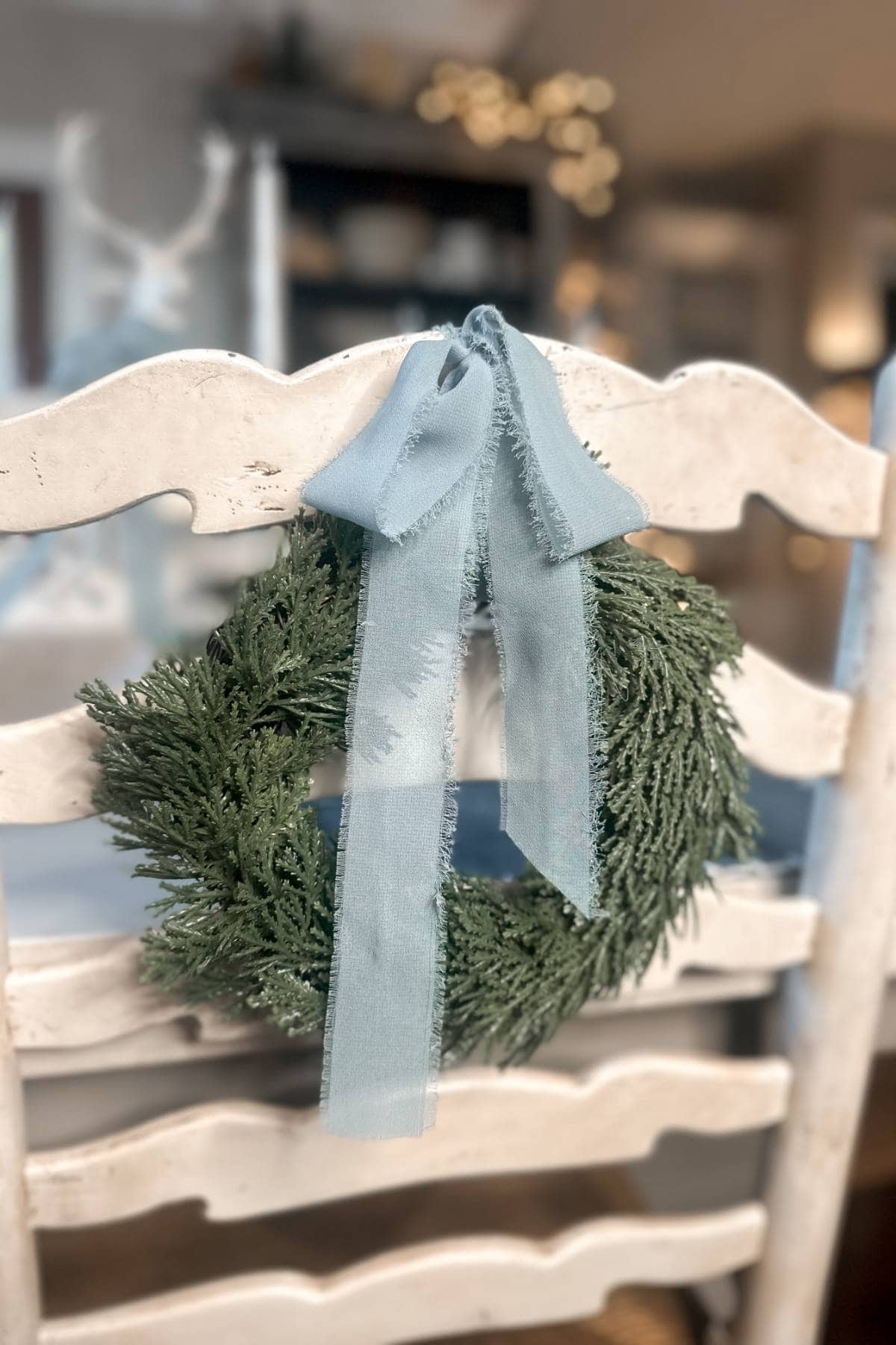 A small green wreath with a light blue ribbon is hanging on the back of a white wooden chair. A softly blurred background features a cozy, warmly lit room.