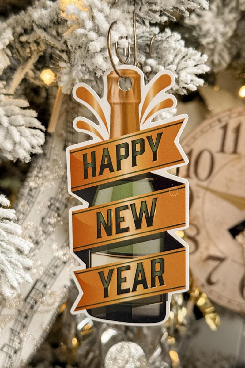 A festive "Happy New Year" ornament depicting a champagne bottle hangs on a snow-covered Christmas tree. The tree is decorated with lights and a large clock face in the background.