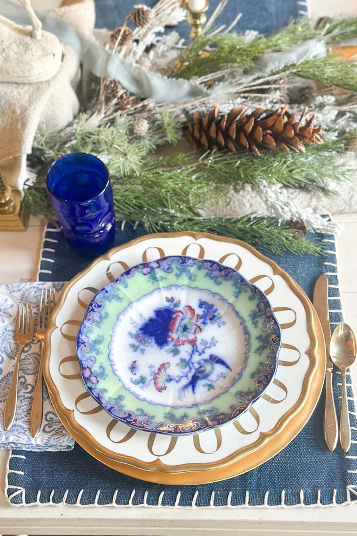 A beautifully set table features a decorative plate with a floral design, a gold-rimmed charger, and blue glass. Silverware and a patterned napkin are placed beside it. Pinecones and greenery adorn the center, suggesting a festive atmosphere.