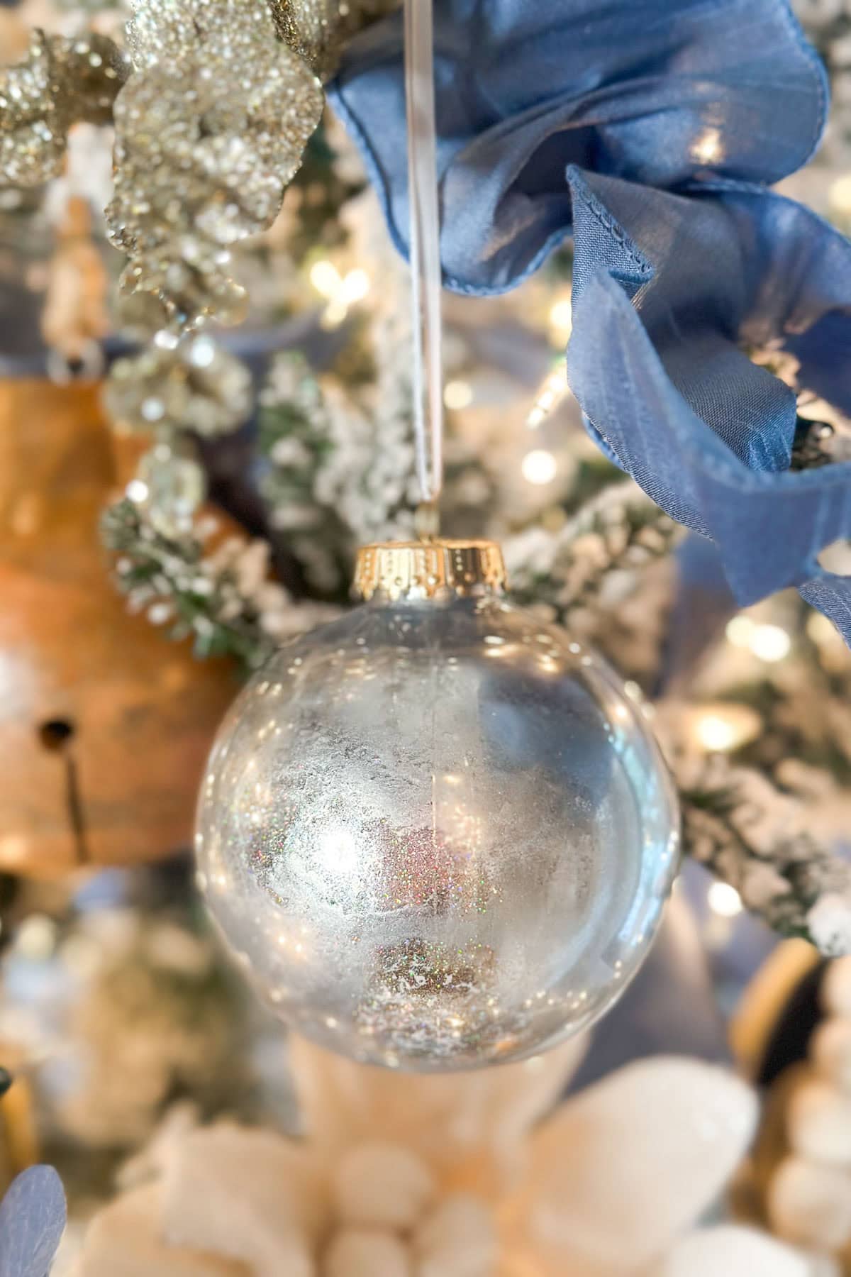 A close-up of a Christmas tree ornament featuring a silver, glittery bauble hanging from a branch. The bauble is surrounded by blue ribbon, golden leaves, and glowing lights, creating a festive and elegant decoration.