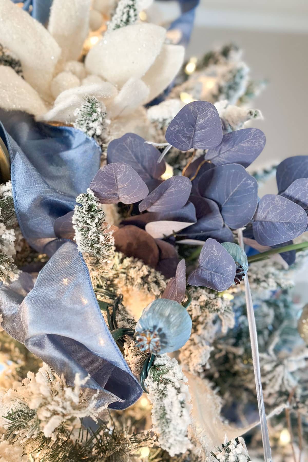 Close-up of a Christmas tree decorated with frosted white flowers, purple leaves, and blue ribbon. Small green ornaments and snow-dusted branches are visible, creating a festive and elegant winter theme.