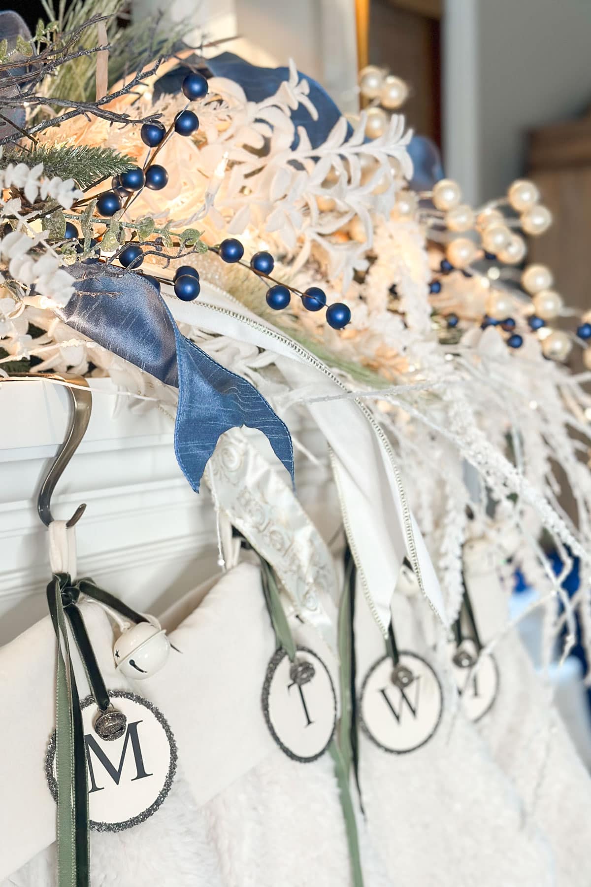 A festive holiday mantel display featuring white and blue decorations, elegant ribbons, and greenery. Stockings hang below, each with a different initial, and a beautifully lit garland drapes across the top.