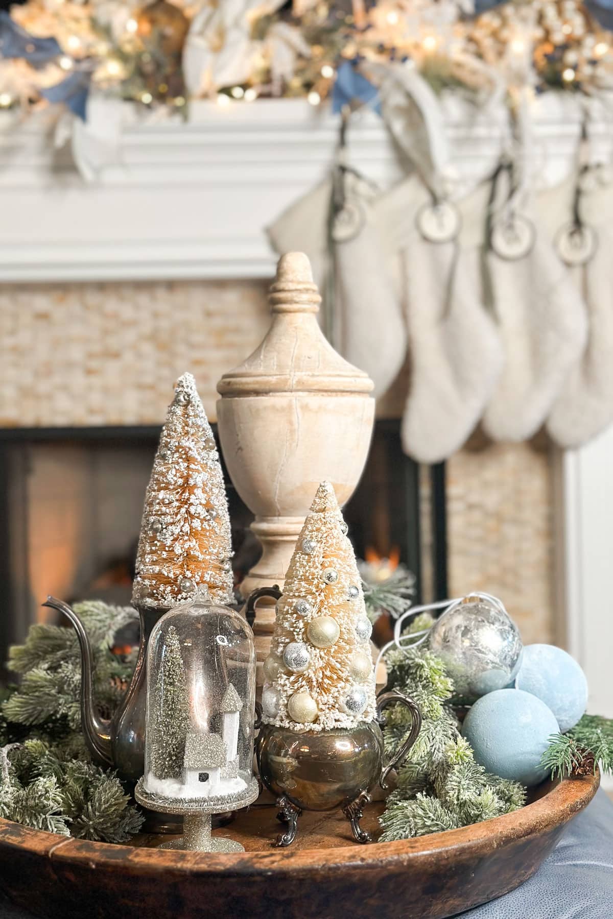 Festive holiday decor featuring cone-shaped trees adorned with snow and ornaments, surrounded by greenery, silver teapots, a snow globe, and large blue ornaments, set on a wooden tray in front of a cozy fireplace and stockings.