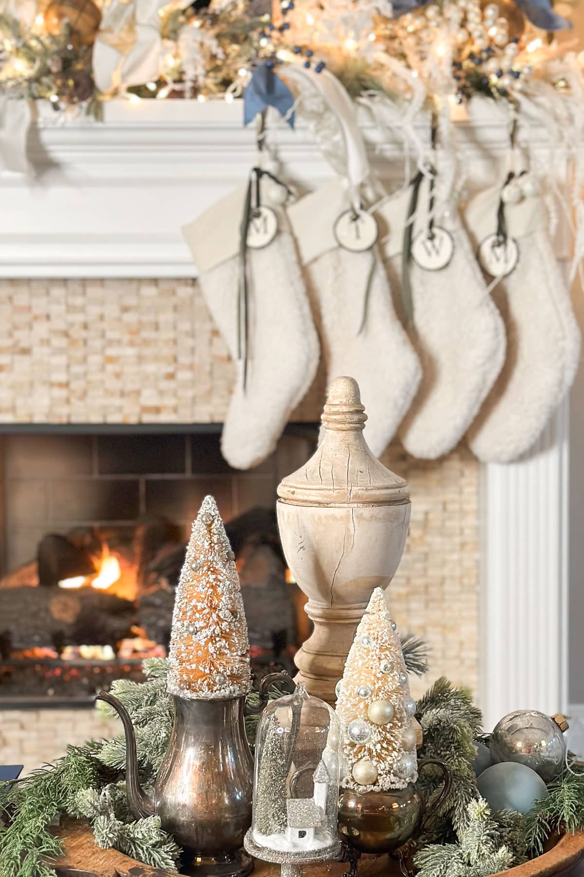 A cozy Christmas scene with a fireplace in the background, decorated with four stockings. The foreground features a table with festive ornaments, including trees made from seashells and a teapot adorned with winter greenery.