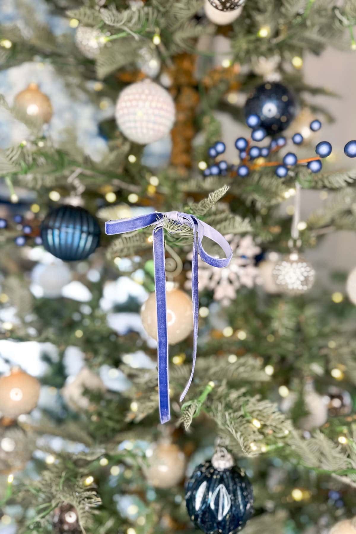 A Christmas tree adorned with blue, white, and metallic ornaments, featuring a prominent blue ribbon in the center. Small lights twinkle amidst the branches, creating a festive and cozy atmosphere.