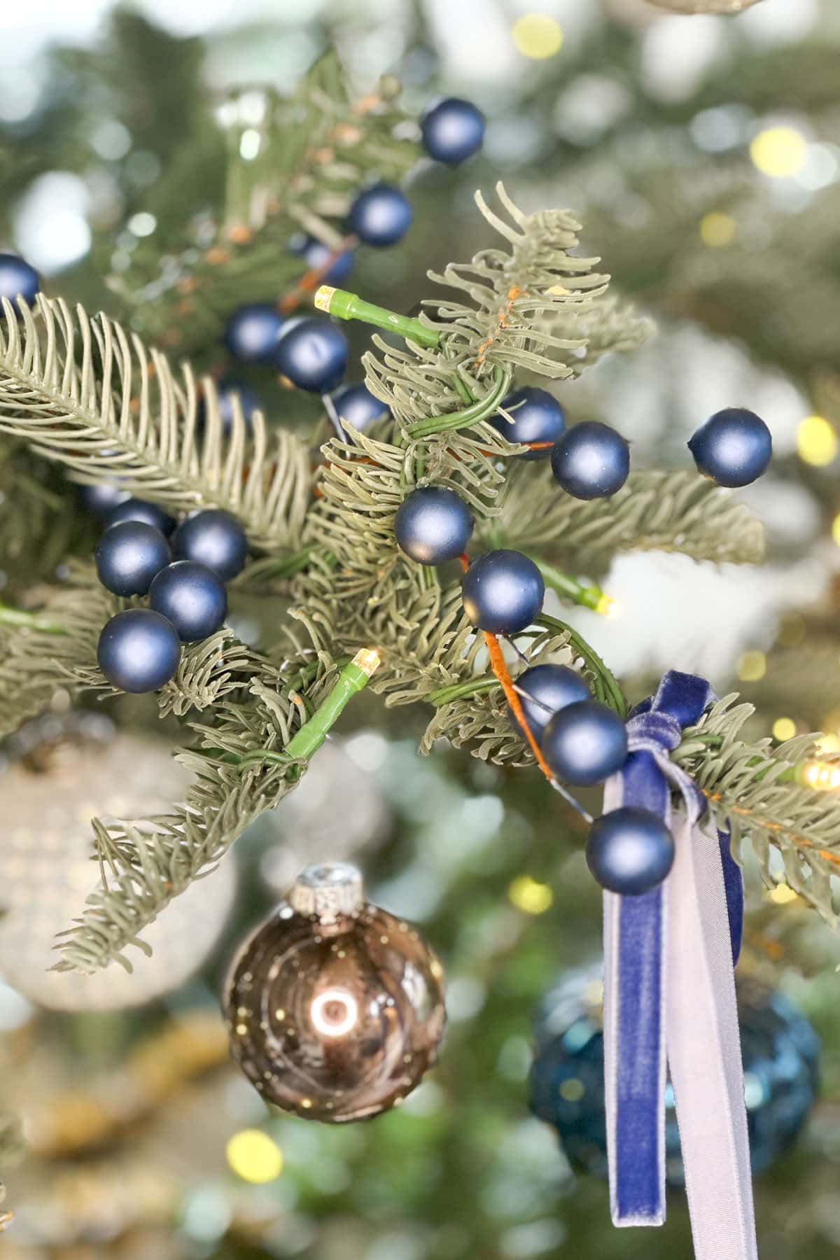 Close-up of a decorated Christmas tree branch featuring blue baubles, a silver ornament, and a blue velvet ribbon. The background is softly blurred, highlighting the festive decorations.