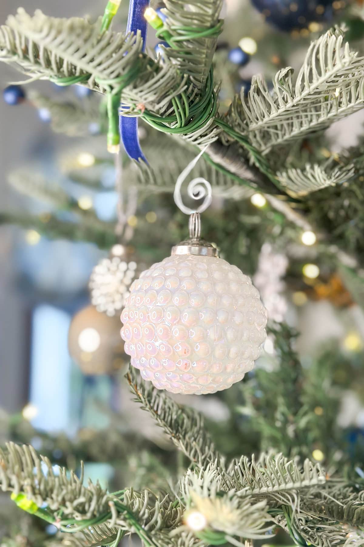 Close-up of a decorated Christmas tree with a white, textured glass ornament hanging from a branch. The tree is adorned with green needles and small, twinkling lights in the background, adding a festive glow.