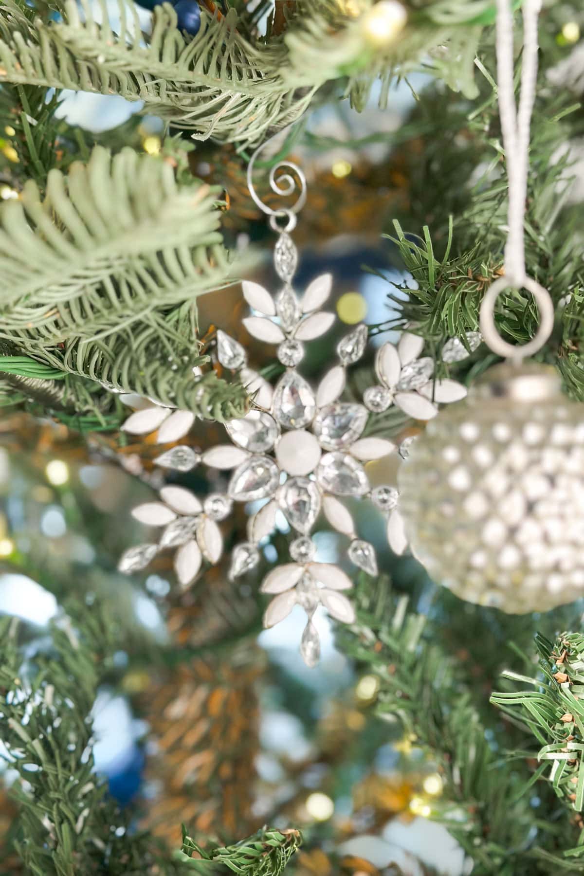 Close-up of a snowflake ornament adorned with crystals, hanging on a Christmas tree. The tree is decorated with green needles and warm lights, accompanied by a textured silver bauble. The background is softly blurred.