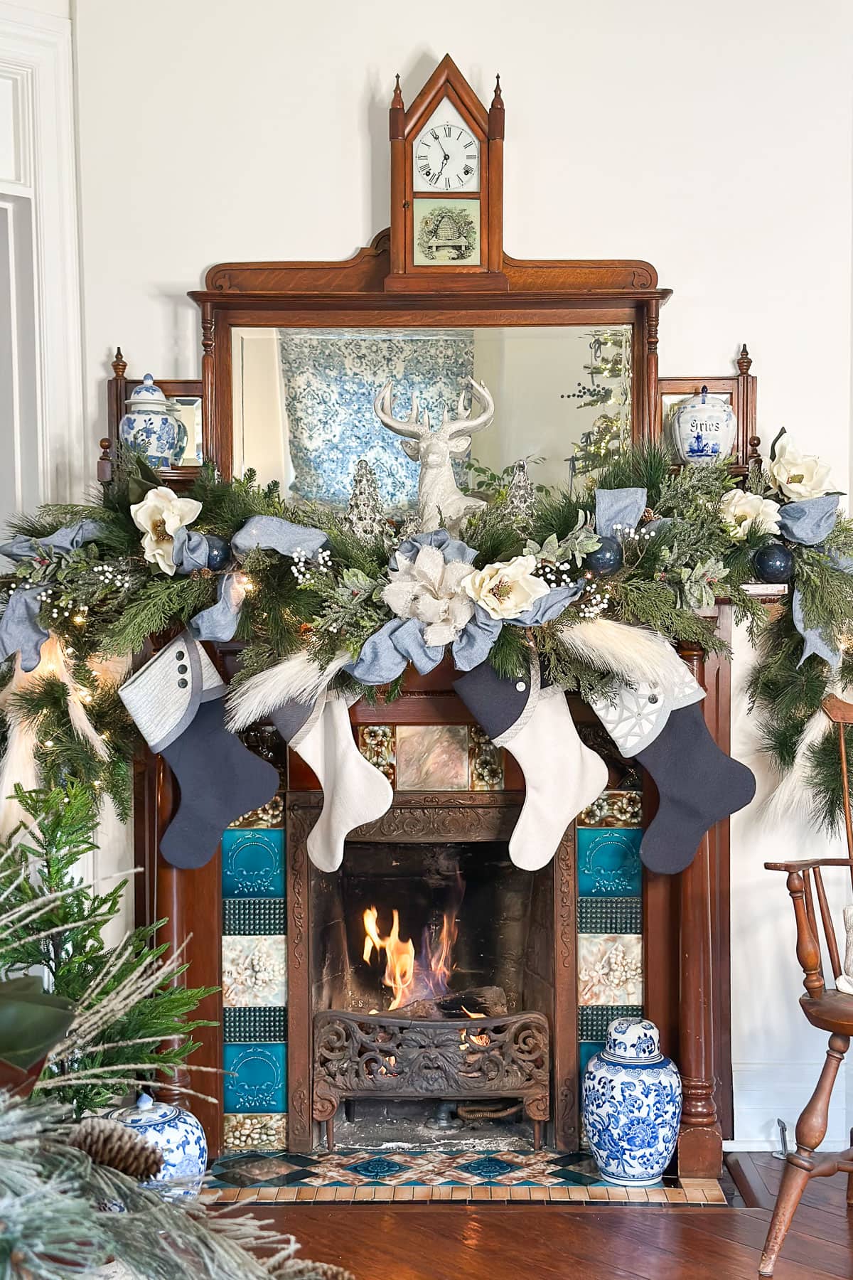 A festive fireplace with a wooden mantel decorated with greenery, white flowers, and ornaments. Four stockings hang below. A fire burns in the hearth, and a clock sits atop the mantel. Bright blue accents and ceramic vases complete the scene.