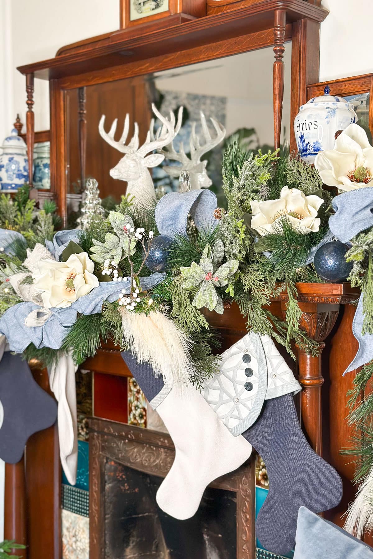 A festive fireplace mantle adorned with greenery, white flowers, and ribbon. A white reindeer figurine is centered. Stockings in shades of gray and white hang below. Blue and white decorative jars are placed on the mantle.