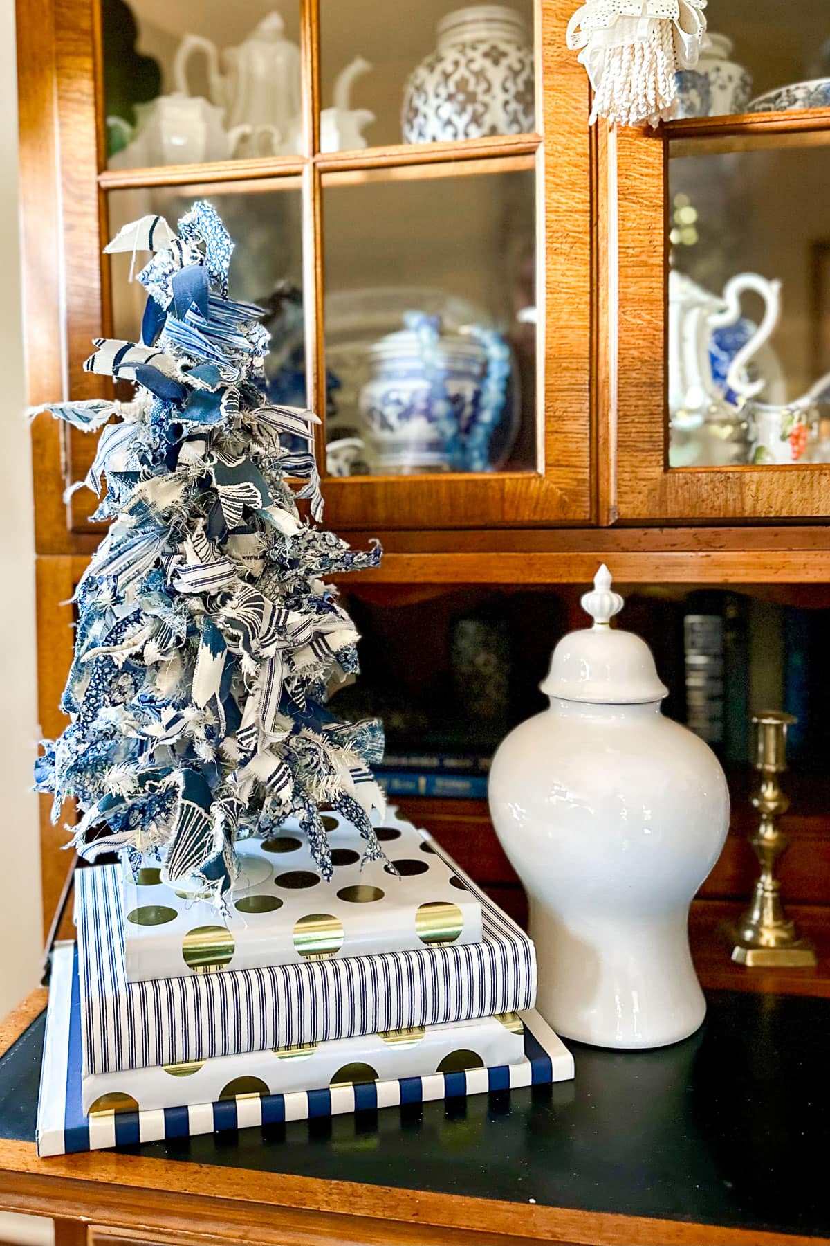 A decorative blue and white fabric Christmas tree stands atop a stack of striped and polka dot boxes. Nearby is a white ceramic jar with a lid. In the background, a wooden cabinet displays blue and white porcelain items.