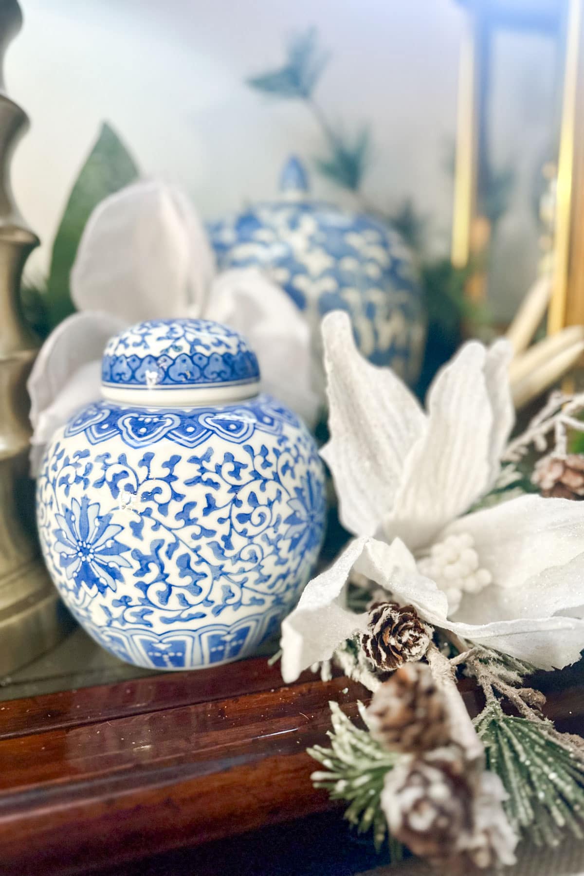 A blue and white ceramic jar with intricate floral patterns sits next to white fabric flowers and pine cones on a wooden surface. The scene is softly lit, giving a cozy and decorative feel.