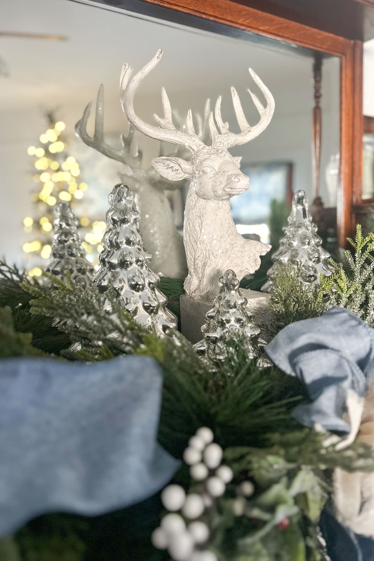 A festive decoration featuring a white reindeer sculpture with large antlers is surrounded by small glass trees, greenery, and blue ribbons. A blurred Christmas tree with lights is in the background.