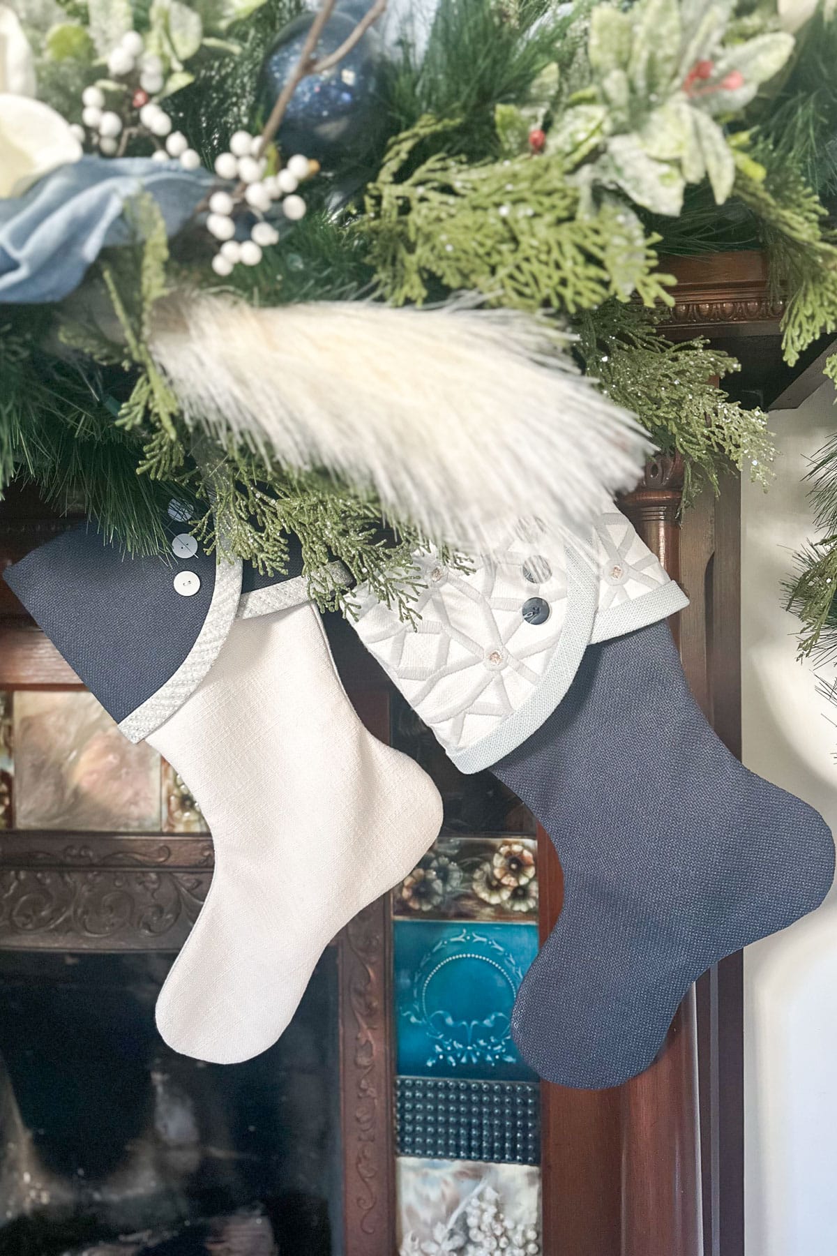 A fireplace decorated for the holidays with two stockings hanging from a mantel adorned with green foliage, white berries, and blue flowers. The stockings are blue and white with button accents.